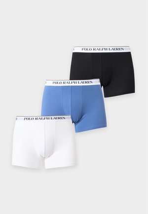 Mladý muž stojaci bosý v čiernych boxerkách s bielym pásom s logom Polo Ralph Lauren na jednofarebnom pozadí.