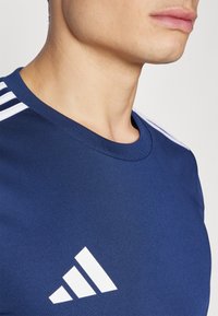 Navy blue athletic t-shirt made of smooth fabric featuring a round neck, white shoulder stripes, and a prominent white logo on the chest.