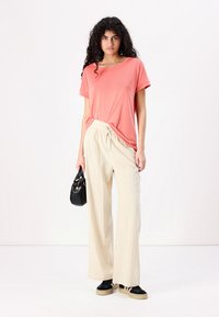 T-shirt corallo a maniche corte, abbinato a pantaloni palazzo beige chiaro e sneaker nere. Una borsa nera aggiunge un tocco di dettaglio all'outfit.