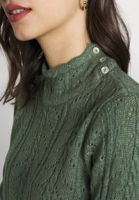 Pull en tricot vert avec un col montant, présentant un motif texturé et trois boutons décoratifs sur l'épaule. Matière douce.