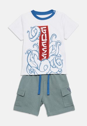 Chemise blanche à manches courtes avec des tentacules de pieuvre bleues et un texte vertical rouge "GUESS", assortie à un short cargo vert avec des cordons bleus.