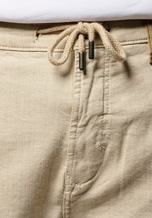 Beige katoenbroeken hebben een tailleband met een trekkoord van gevlochten touw en metalen uiteinden, een vijf-pocket ontwerp en een subtiele textuur in de stof.