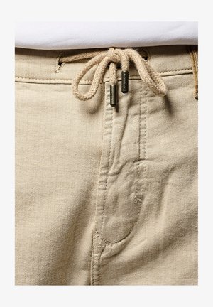 Los pantalones de algodón beige presentan una cintura con cordón ajustable de cuerda trenzada y puntas metálicas, un diseño de cinco bolsillos y una textura sutil en la tela.
