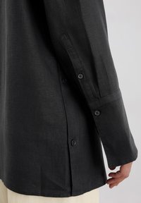 Schwarzes Langarmshirt aus strukturiertem Stoff, mit Knöpfen an den Manschetten und einem geschwungenen Saum mit seitlichen Schlitzen.