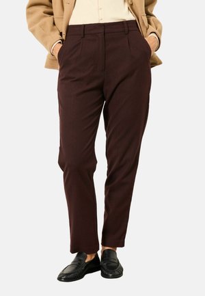 Person wearing dark brown tailored pants with pockets, black loafers, a beige jacket, and a light shirt, standing with hands in pockets.