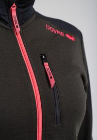 Mørkegrøn zip-up jakke med rosa og sorte accenter. Har en frontlynlås og logo. Glat tekstur og tætsiddende design.