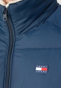 Mörkblå puffjacka med hög krage, med dragkedjestängning och en texturerad innerväv. Har ett rektangulärt logopatch.