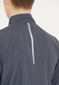 Merijacketti korkealla kauluksella, jossa on heijastava pystysuora raita takana ja sileä, kevyt kankaan pinta.