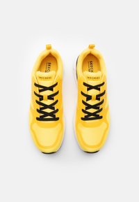 Yellow athletic shoes with a textured design, black laces, and a reinforced toe. Branding visible on the tongue and side detailing.