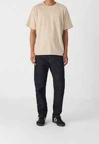 Beige bomulls t-shirt med korta ärmar, rund halsringning och subtil logotyp. Matchad med mörkblå denimjeans och svarta sneakers med vita ränder.