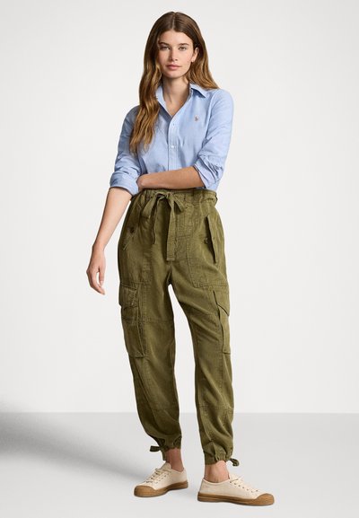 Γυναίκα που φοράει ανοιχτό μπλε πουκάμισο με κουμπιά και χακί παντελόνι cargo με ζώνη στη μέση και μπεζ αθλητικά παπούτσια, στέκεται μπροστά σε απλό φόντο.