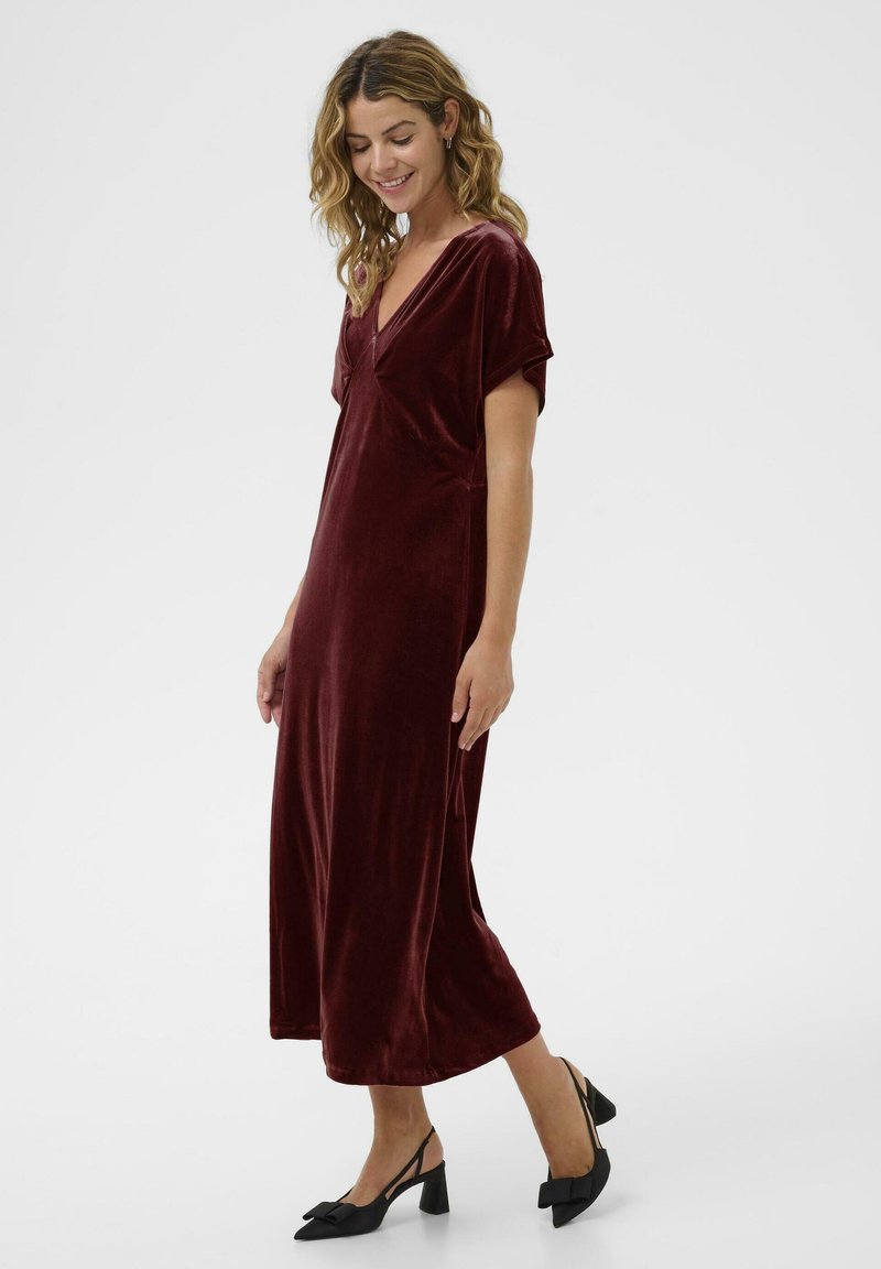 Kvinna som står och ler, iklädd en vinröd velour midi-klänning med korta ärmar och svarta låga klackar med rosetter.