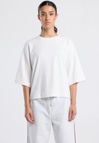 Woman with dark pulled-back hair wearing a loose white short-sleeve top and light gray sweatpants against a plain background.