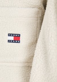 Crèmekleurige fleece jas met een vierkante patch met de tekst "TOMMY JEANS" en een kleurblokontwerp in blauw, rood en wit.