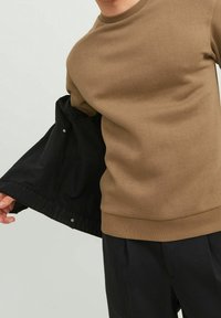 Sudadera marrón con cuello redondo y puños acanalados, combinada con una chaqueta negra con cierre a presión en la manga.