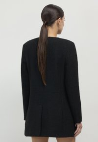 Blazer nero con texture, taglio dritto, maniche lunghe e dettagli di apertura sul retro. Vista anteriore non mostrata, focus sul design posteriore.