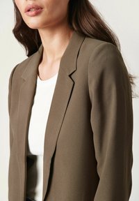 Olivově zelený blazer s jednobarevným designem, klopovými límci a hladkou texturou, nošený přes bílý žebrovaný top.