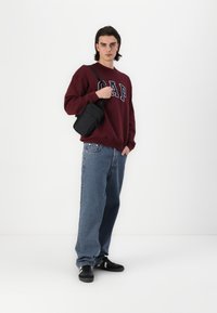 Vinröd sweatshirt med "GAP"-logotyp, blå jeans och svart axelväska. Svarta sneakers med vita ränder. Enkelt, avslappnat passformdesign.