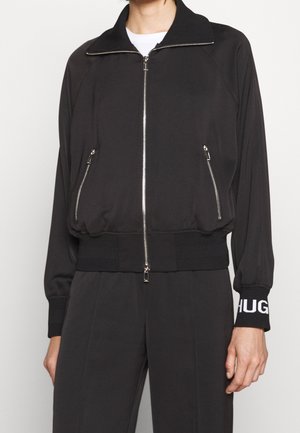 Personne portant une veste noire zippée avec des fermetures éclair argentées sur les poches et les poignets, montrant des lettres blanches partielles sur le poignet droit.