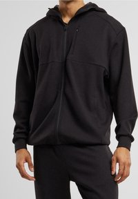 Sudadera con cremallera negra hecha de tejido suave, con capucha, bolsillos laterales y un panel texturizado en el pecho. Diseño simplista sin logotipos.