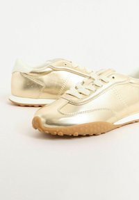 Zlaté metalické tenisky s hladkým povrchem, bílými akcenty a gumovou podrážkou s texturovaným gripem. Šněrovací design s jasně viditelnými švy.