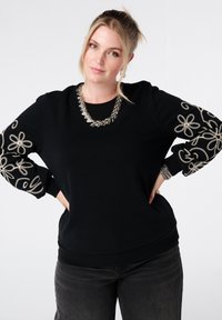 Zwarte sweatshirt met geborduurde zilveren bloem- en spiraalpatronen op de mouwen. Heeft een ronde halslijn en een geribbelde onderrand.