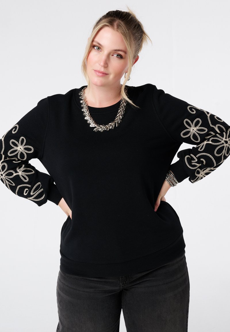 Zwarte sweatshirt met geborduurde zilveren bloem- en spiraalpatronen op de mouwen. Heeft een ronde halslijn en een geribbelde onderrand.