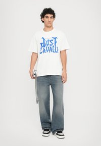 White cotton t-shirt featuring large blue "Just Cavalli" graphic, relaxed fit. Paired with lightly distressed blue jeans and black/white sneakers.
