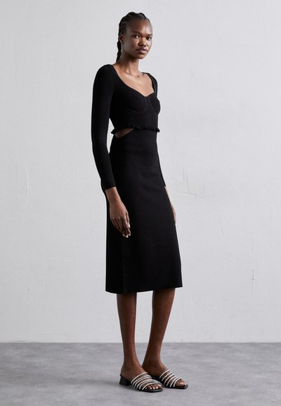 Schwarzes Strickkleid mit langen Ärmeln, gerafftem Taillenbund und Ausschnitten; kombiniert mit schwarzen Rutschsandalen, verziert mit weißen Perlen.
