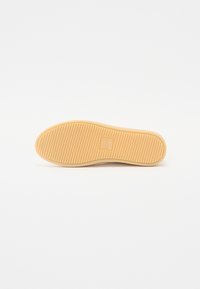 Tan rubber shoe sole with horizontal ridges for grip, featuring a small embossed logo in a square near the heel. Smooth texture overall.