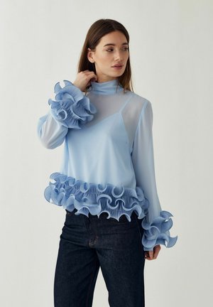 Kobieta ubrana w jasnoniebieską, przezroczystą bluzkę z falbanami na mankietach i dole, do tego ciemne dżinsy, stoi na tle jednolitego jasnego tła.