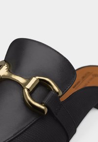 Mocassin en cuir noir avec sangle texturée et détail en métal doré en forme de mors de cheval sur le dessus, sur un fond uni.