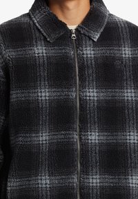 Black and gray fleece jacket with a checked pattern, featuring a zip closure and a pointed collar. Soft texture and warm material.
