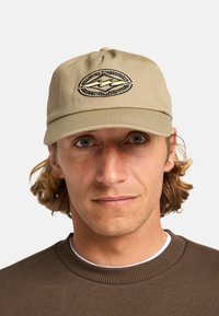 Beige baseball cap made of cotton with a curved brim. Features an embroidered logo in dark thread. Simple, structured design.