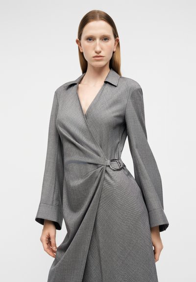 Γυναίκα με ίσια μαλλιά που φοράει ένα γκρι φόρεμα με μακριά μανίκια και ζώνη με μεταλλική αγκράφα στη δεξιά πλευρά, μπροστά σε απλό φόντο.