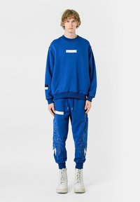 Sudadera de algodón azul con una etiqueta en el pecho, combinada con pantalones de chándal a juego que presentan bordados azules parecidos a plumas y acentos blancos.