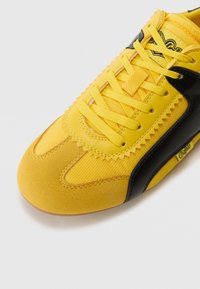 Sárga sportcipő fekete kiegészítőkkel, textil és velúr anyagok felhasználásával, texturált felülettel, éles varrással és lapos fűzővel.