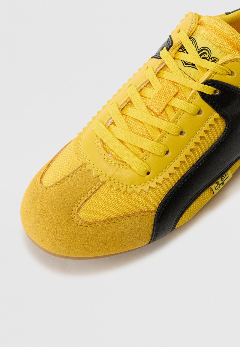 Zapatilla amarilla con acentos negros, que presenta materiales de textil y ante, superficie texturizada, costuras definidas y cordones planos.