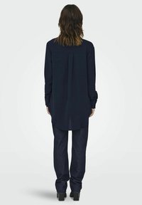 Navy long-sleeve shirt with a straight cut, back pleat, rounded hem, and buttoned cuffs, paired with dark denim trousers and heeled shoes.