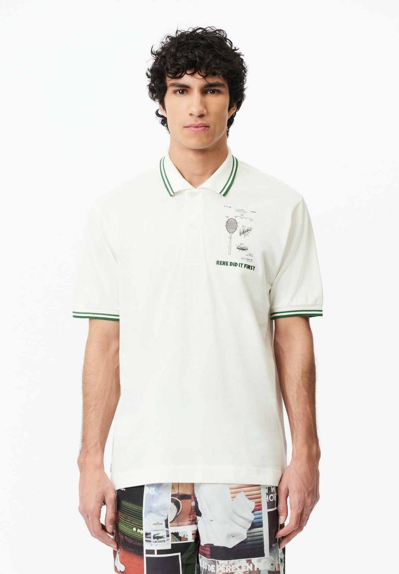 Polo shirt blanc avec col et rayures vertes aux manches. Présente un motif imprimé et un texte sur la zone gauche de la poitrine. Texture de tissu douce.