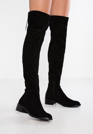 Zwarte suède over-de-knie laarzen met lage blokhakken, gedragen door een persoon die staat op een witte vloer tegen een effen achtergrond.