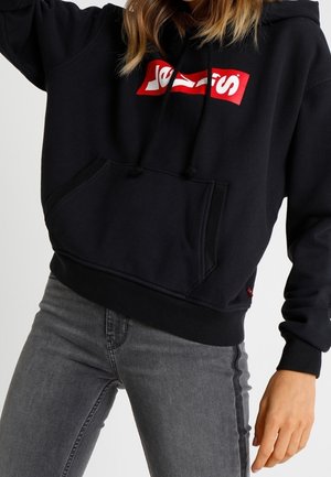 Femme portant un sweat à capuche noir avec un texte graphique rouge et blanc, assorti à un jean gris, debout devant un fond uni.