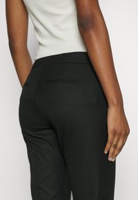 Tylny widok osoby noszącej czarne dopasowane spodnie z rozciętymi tylnymi kieszeniami oraz białą bezrekawnikową bluzkę.