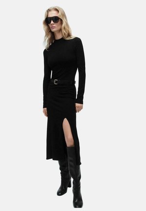Robe noire à manches longues avec une fente sur le côté et une taille ceinturée. Associe à des bottes noires montantes et des lunettes de soleil noires surdimensionnées.