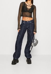 Svart nät långärmad crop-top med nyckelhålsknäppning, kombinerad med mörkblå vida denimcargobyxor med vit sömnad och fickor. Håller en silverfärgad metallisk mini-handväska.