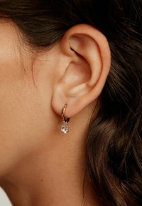 Aro de oro rosa con una gema rectangular clara colgando de él. El aro se muestra usado en la oreja, destacando su diseño.