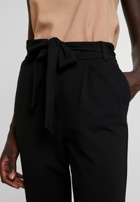 Svarta skräddarsydda byxor med en bred midjeband, som har en tygrem. Slät, mjuk textur, sidofickor och en figurnära silhuett.