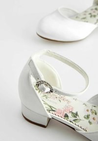 Pantofi albi din satin cu bretea la gleznă și toc mic, cu brant floral și o cataramă decorativă din argint pe bretea.