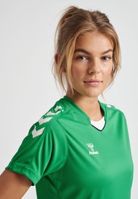 Grøn sportstrøje med v-hals, korte ærmer og hvide detaljer. Lavet af glat stof med et logo på forsiden.