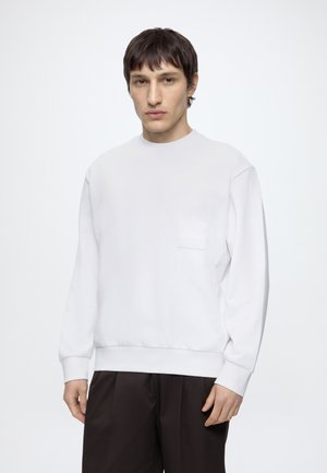 Jeune homme aux cheveux courts et foncés portant un sweat-shirt blanc à manches longues et un pantalon noir, debout devant un fond clair uni.